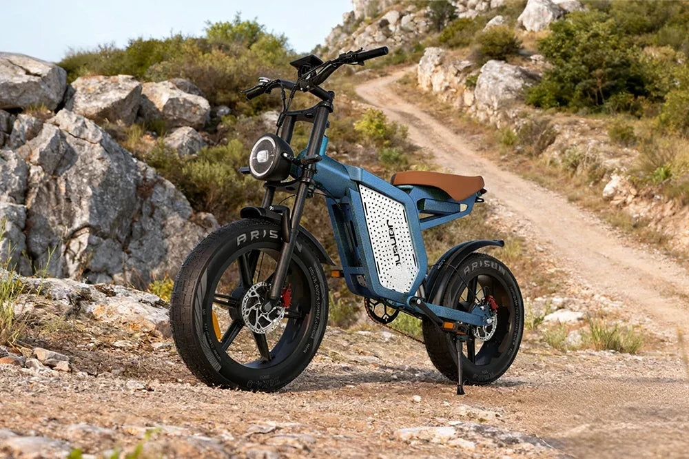 Stylish blue Jansno X60 retro electric bike parked on rocky mountain dirt trail amid rugged hillside scenery.