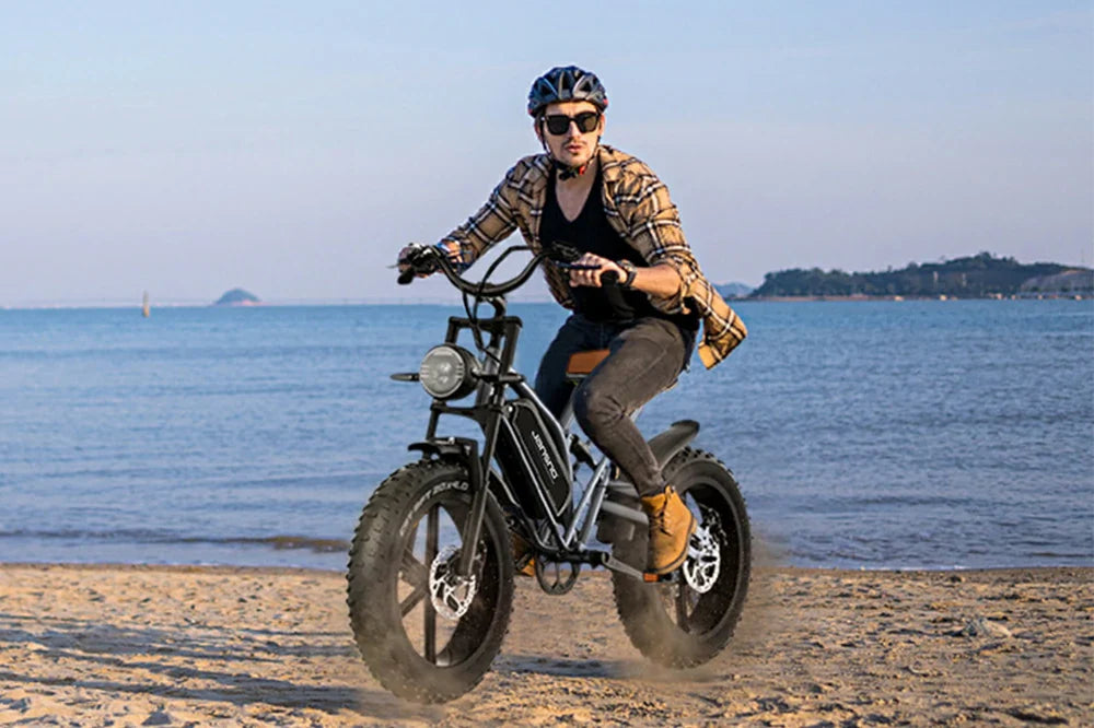Black vintage-style X50 all-terrain electric bike with rugged thick fat tyres, man in plaid shirt riding across coastal sand beside calm blue seawater and distant islands