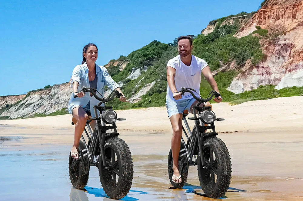 Matte black Jansno X50 vintage-style electric bikes with oversized wide sand-proof fat tyres and round front headlights, man and woman cruising shallow shore water beside layered colourful coastal cliffs and bright blue sky