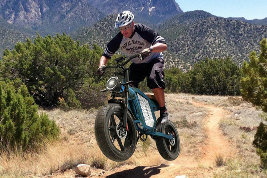 Teal metallic Jansno X60 vintage-style electric cruiser bike, male rider lifting front wheel while off-roading on dry dirt path surrounded by dense shrubs and rugged rocky mountain ranges