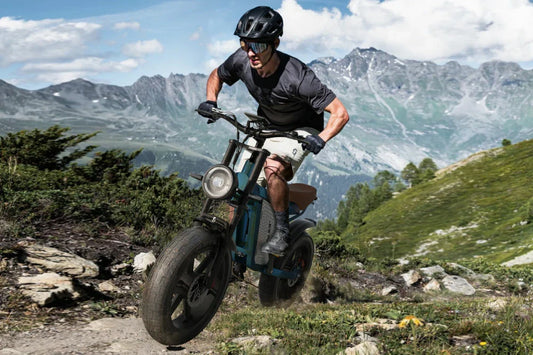 Dynamic mountain trail action shot of rider navigating teal Jansno X60 retro fat tire electric bike over rocky alpine terrain with snow-capped peaks backdrop.
