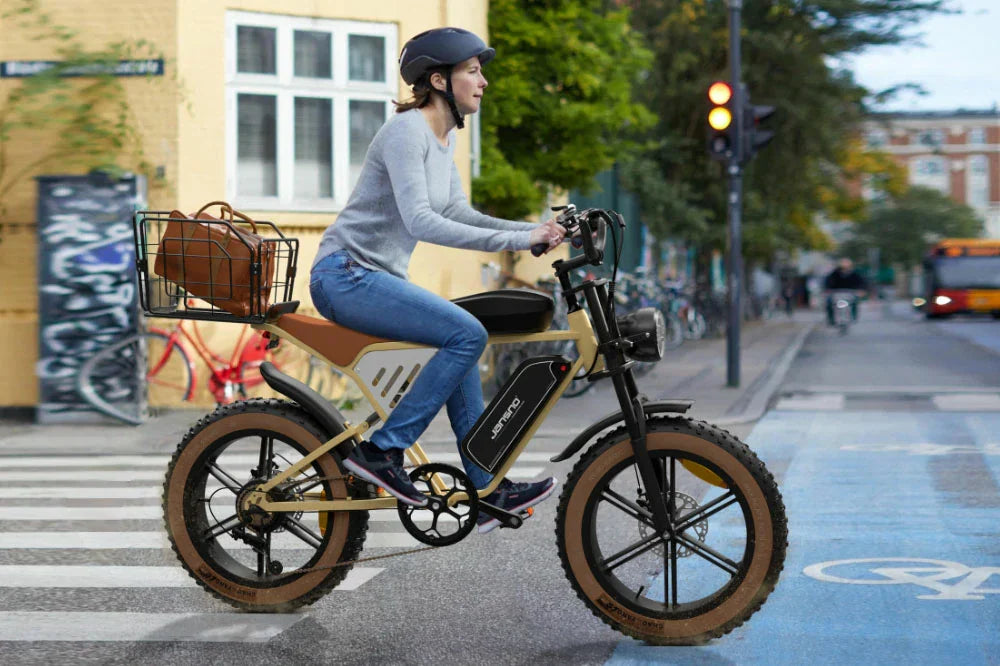 Urban commute lifestyle shot of a helmeted female rider cruising cream retro Jansno X70 e-bike with rear cargo basket across city crosswalk at a street intersection.