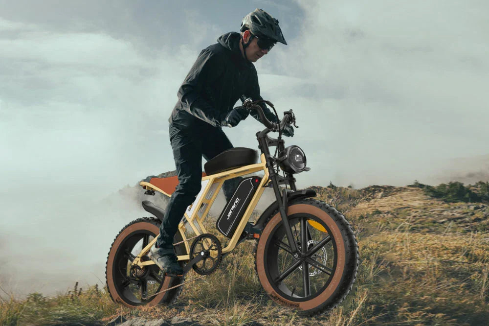 Dynamic misty hillside action shot of helmeted male rider traversing rugged grassy highland trails on cream retro-styled Jansno X70 fat tire electric bike.